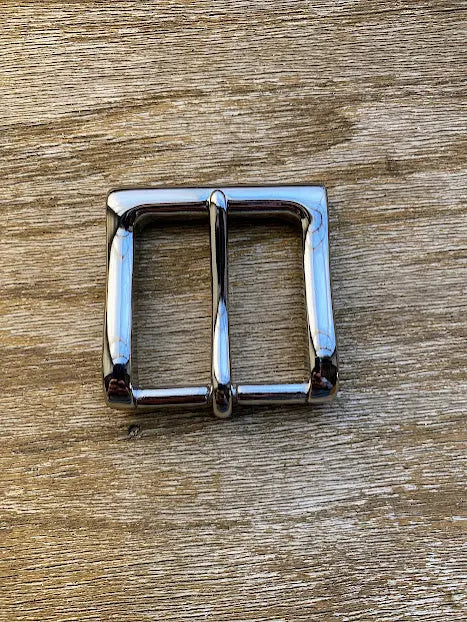 A shiny, rectangular silver buckle sits on a textured wooden surface, ideal for pairing with the BBH Handcrafted Leather Belt.