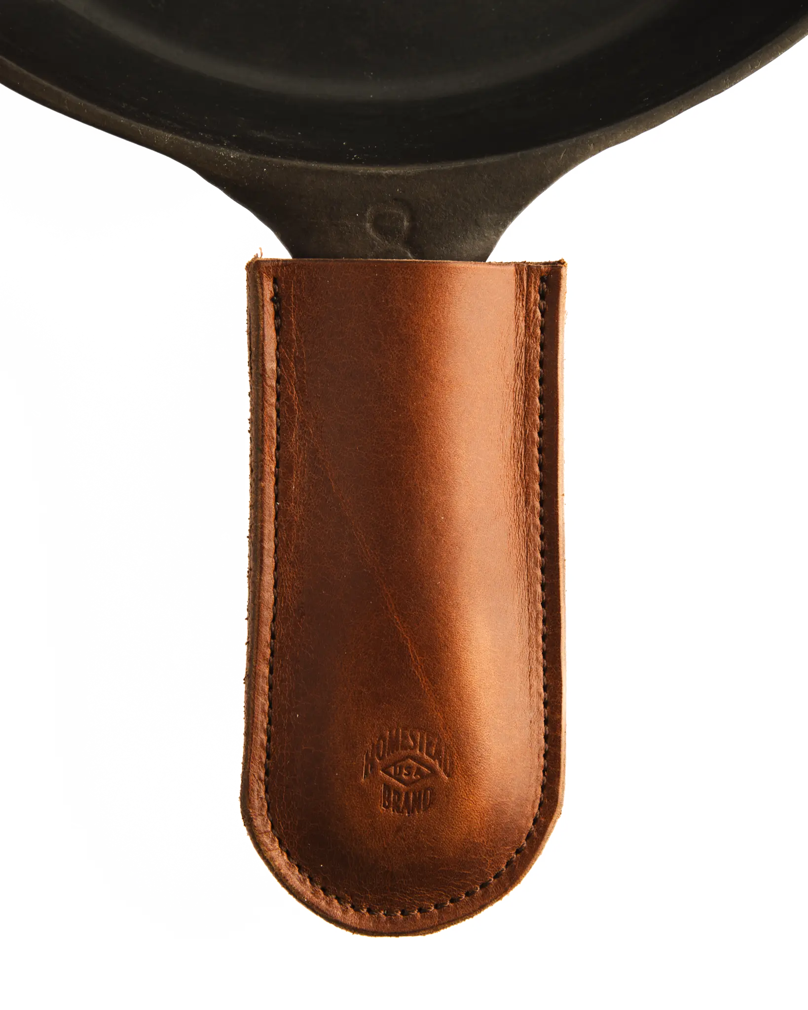 The Homestead Brand Skillet Handle Sleeve No. 2002, made of brown full grain leather with visible stitching and a debossed logo, slips over the handle of a black cast iron skillet.