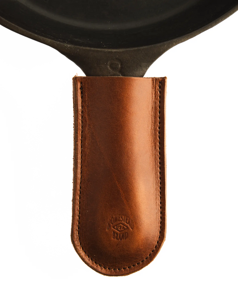 The Homestead Brand Skillet Handle Sleeve No. 2002, made of brown full grain leather with visible stitching and a debossed logo, slips over the handle of a black cast iron skillet.