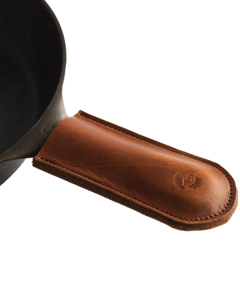 A close-up of the Homestead Brand Skillet Handle Sleeve No. 2002, a brown full grain leather cover stitched at the edges and embossed with a small logo near its rounded end, fitted on a cast iron skillet handle.