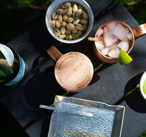Luxury Drink Stirrers by Havens | Luxury Metals, along with two copper mugs (one with ice and lime), a bowl of pistachios, a bottle, and a textured tray, are arranged on a dark outdoor table in sunlight.