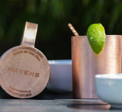 A Havens | Luxury Metals copper mug with HAVENS engraved on the bottom lies beside another filled mug, garnished with a lime wedge and Luxury Drink Stirrer; white bowls are blurred in the foreground.