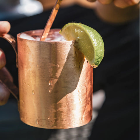 A hand holds a copper mug with a frothy drink and lime wedge, featuring a Havens | Luxury Metals Luxury Drink Stirrer inside. The background is softly blurred.