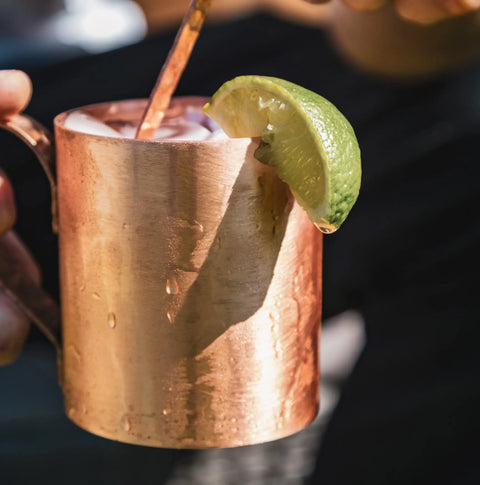 A person holds a copper mug with a lime wedge and cold drink, featuring condensation and a metal stirrer from Havens | Luxury Metals Premium Metal Drink Stirrers—handcrafted bar accessories made in the USA.