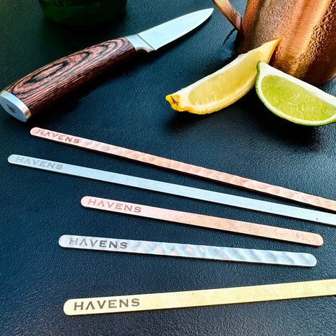 A knife with a wooden handle, a copper cup, lemon and lime wedges, and five Havens | Luxury Metals Premium Metal Drink Stirrers – Handcrafted Bar Accessories (Made in USA)—ideal for cocktail décor—are arranged on a dark surface.