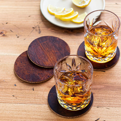 Two glasses of amber whiskey sit on the Moose Leather Coaster Set by Rogue Industries atop a wooden table. In the background, a white plate with lemon wedges adds a fresh touch to the scene.