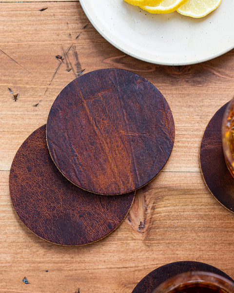 Two round, brown coasters from the Moose Leather Coaster Set by Rogue Industries sit on a wooden table, alongside a white plate with lemon slices and a beverage glass.