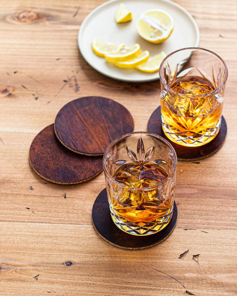 Two crystal glasses of amber whiskey rest on the Rogue Industries Moose Leather Coaster Set atop a wooden table. In the background, a plate displays sliced lemons—some as wedges, others as halves.