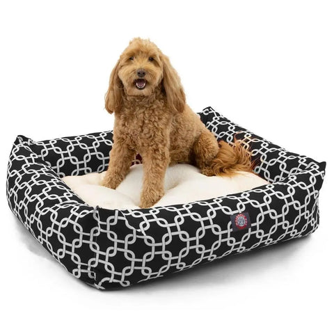 A curly-haired, light brown dog sits happily on the Majestic Pet Links Luxe Bagel Bed, a plush black and white patterned bed with raised sides.