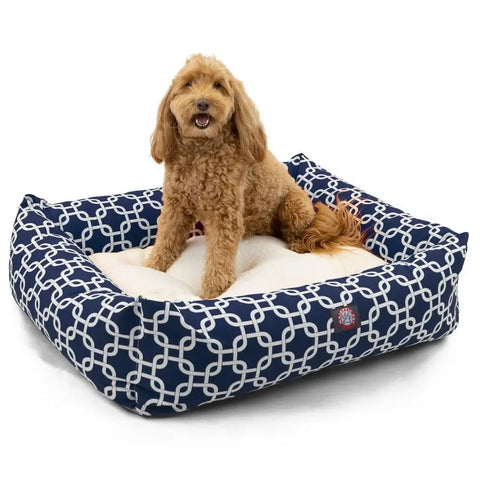 A fluffy, light brown dog sits happily on the Majestic Pet Links Luxe Bagel Bed, a plush navy blue bed with a white geometric pattern, looking forward with its mouth open as if smiling.