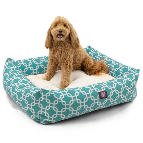 A small, curly-haired brown dog sits on a Majestic Pet Links Luxe Bagel Bed with teal and white patterns and cream-colored cushioning, looking forward with its mouth open.