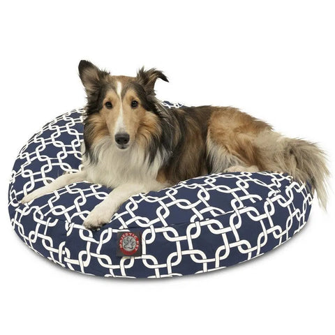 A long-haired dog with brown, black, and white fur is lying on the Majestic Pet Links Round Dog Bed, which features a navy blue and white geometric pattern and a removable cover. The dog is looking toward the camera.
