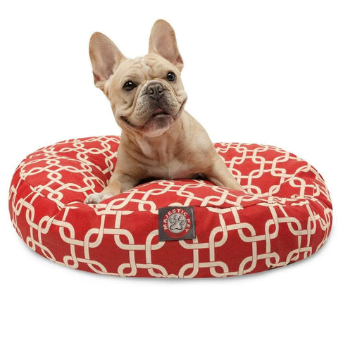 A light brown French Bulldog sits on a Majestic Pet Links Round Dog Bed, which is red with a white geometric pattern, has a removable cover, and displays the brand tag on the front.