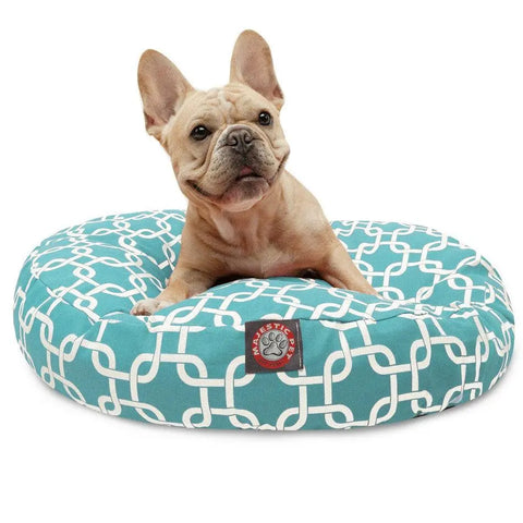 A light brown French Bulldog sits on a teal Majestic Pet Links Round Dog Bed with a white geometric pattern and red brand tag. The bed features a removable, washable cover. The dog gazes up with perked ears.