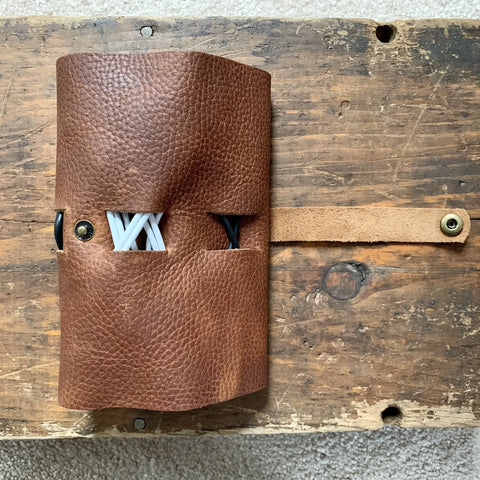The Rogue Industries Leather Tech Organizer, crafted from brown top-grain leather, holds cables and cord holders with white cord ends visible. It sits on a rustic wooden surface, its tan strap extended to the side.