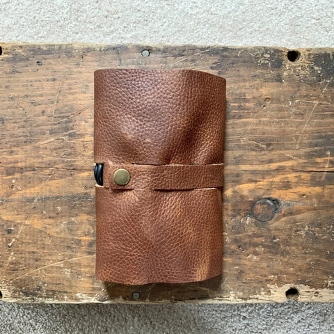 The Rogue Industries Leather Tech Organizer, crafted from brown top-grain leather with a strap and snap button closure, rests on a rustic wooden surface—an ideal solution for stylishly storing cords and devices.