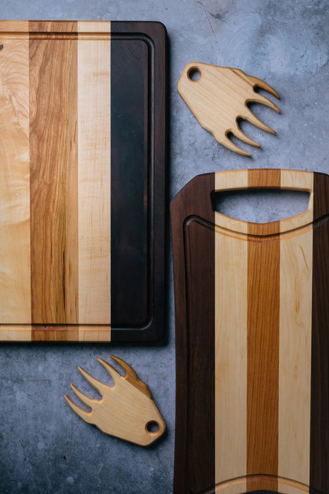 Two wooden cutting boards and two meat claws from the Montana Block Grill Master bundle are displayed on a gray surface. The boards show alternating light and dark wood patterns, with the BBQ tools set near each one.
