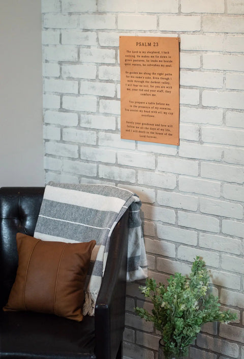 A black leather chair with a brown pillow and striped blanket sits by a green plant. Above, the Urban Southern Psalm 23 Leather Sign is displayed on the white brick wall.