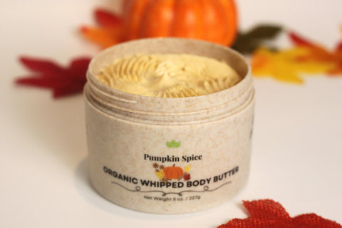An open jar of Forev'Herbs Pumpkin Spice Organic Whipped Body Butter sits on a white surface amid autumn leaves and a decorative pumpkin, inviting deep hydration with its rich, whipped organic texture.