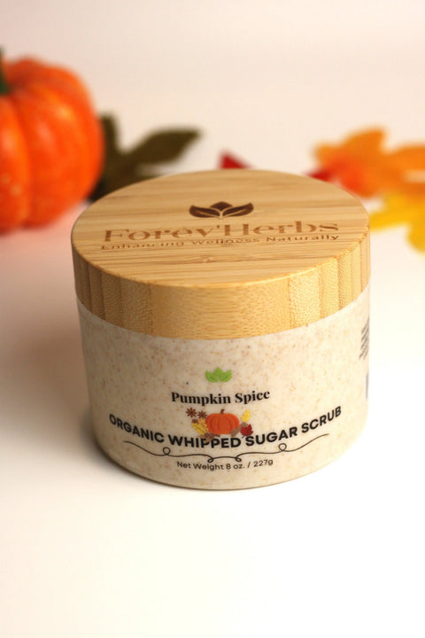 A jar of Forev'Herbs Pumpkin Spice Organic Whipped Sugar Scrub with a wooden lid rests on a white surface, surrounded by blurred fall leaves and a small pumpkin, creating cozy autumn vibes.