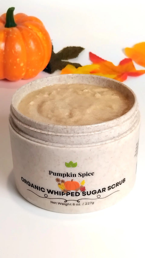 An open jar of Forev'Herbs Pumpkin Spice Organic Whipped Sugar Scrub sits beside a small pumpkin and autumn leaves on a white surface, offering a creamy, textured exfoliation for a perfect touch of fall indulgence.