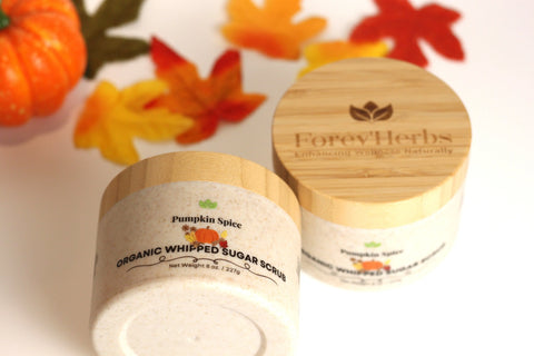 Two jars of Forev'Herbs Pumpkin Spice Organic Whipped Sugar Scrub rest on a white surface, surrounded by autumn leaves and a small decorative pumpkin.