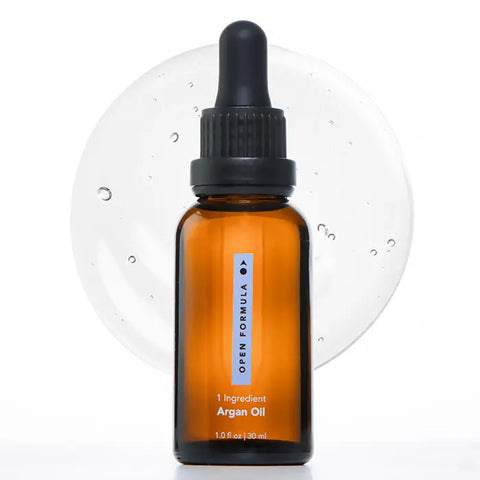 A brown glass dropper bottle labeled Open Formula Argan Oil stands before a clear liquid drop with bubbles on a white background—ideal for those seeking skin hydration and frizz control.