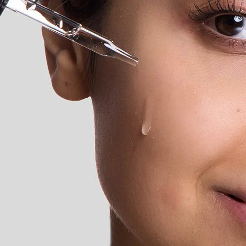Close-up of a womans face as a dropper applies Open Formula Argan Oil serum to her cheek. Her skin looks smooth and healthy, with only one eye, part of her nose, and a hint of a smile showing.