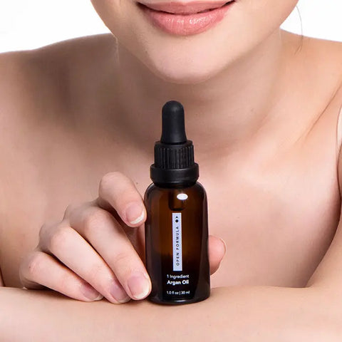A person with bare shoulders smiles gently while holding an Open Formula Argan Oil dropper bottle near their face—this minimalist product tames frizz and moisturizes skin for a healthy glow.