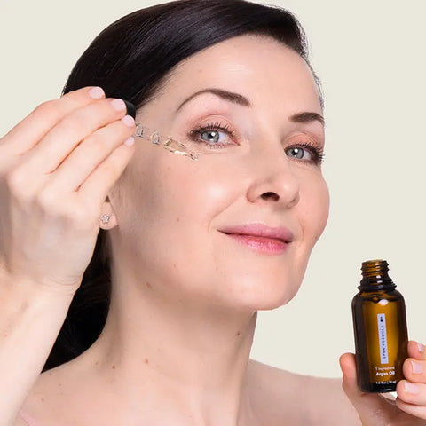 A woman with fair skin smiles at the camera while applying Open Formula Argan Oil facial serum from a dropper bottle to her cheek, moisturizing her skin.
