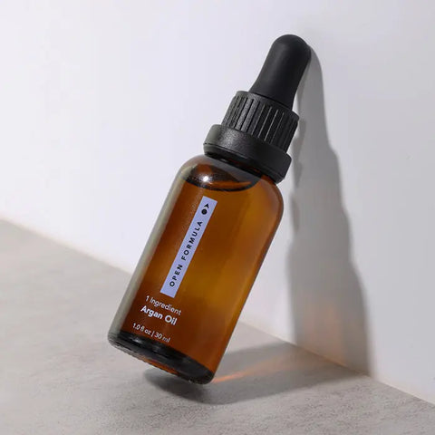 A brown glass dropper bottle labeled Open Formula Argan Oil rests on a light gray surface, casting a soft shadow. This nourishing oil moisturizes skin and tames frizz, leaving you with healthy, radiant results.