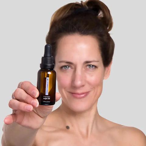 A woman with light skin and brown hair smiles softly, holding a small amber dropper bottle of Open Formula Argan Oil, which moisturizes skin and tames frizz, against a plain gray background.