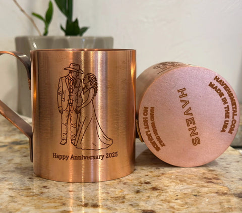 A Pure Copper Mule Mug by Havens Luxury Metals, engraved with a wedding couple and Happy Anniversary 2025, sits on the counter. The base reads HAVENS and handcrafted in USA. A blurred plant appears in the background.