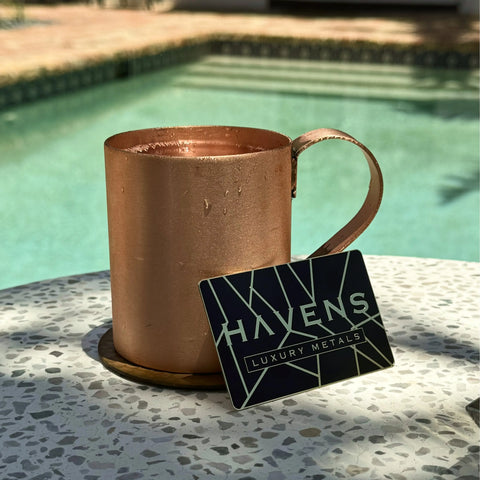 A Pure Copper Mule Mug by Havens Luxury Metals, made in the USA, sits with visible condensation on a white speckled table near a pool beside a black and gold Havens Luxury Metals card, adding elegance to any setting.