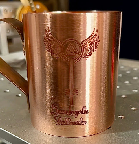 The Pure Copper Mule Mug by Havens Luxury Metals features a winged emblem above Queen Layartha Shieldmaiden. Handcrafted in the USA, this premium mug is shown resting on metal with shelves softly blurred in the background.