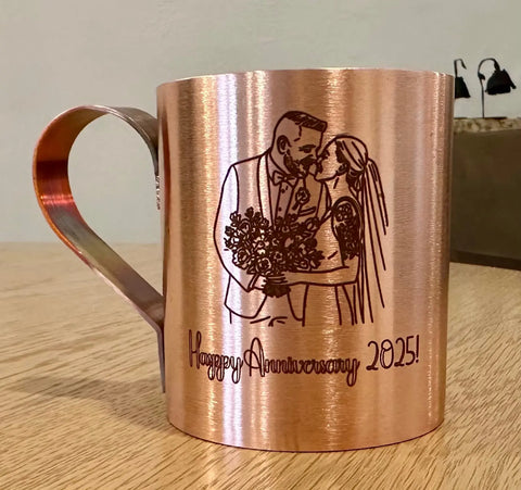 The Havens Luxury Metals Pure Copper Mule Mug, handcrafted in the USA, features an engraved kissing couple in wedding attire with a bouquet and Happy Anniversary 2025! at the bottom. The mug is pictured on a wooden surface.