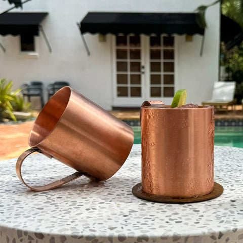 Two Havens Luxury Metals Pure Copper Mule Mugs—USA Made—rest on a speckled outdoor table by the pool; one mug stands upright with a lime garnish while the other lies on its side. A white building with black awnings is in the background.