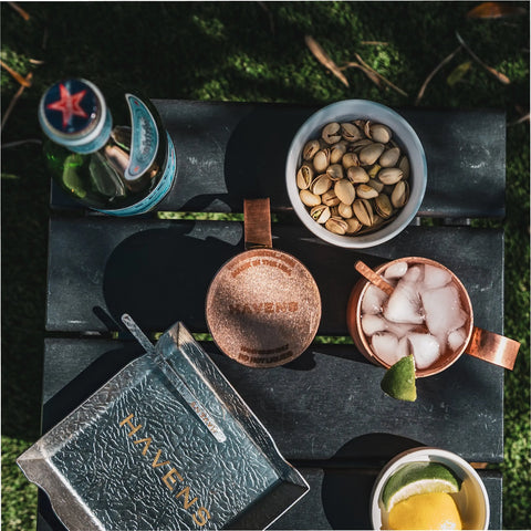 An outdoor tabletop displays a Pure Copper Mule Mug (USA Made) by Havens Luxury Metals with ice and lime, pistachios, a drink bottle, lemon wedges, and a silver tray labeled HAVENS, all sunlit.