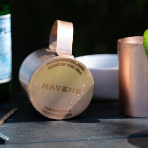 A Pure Copper Mule Mug by Havens Luxury Metals, USA made and engraved with HAVENS, lies on its side on a dark surface next to a whole lime, copper cup, white bowl, and green glass bottle. Sunlight highlights the mug.