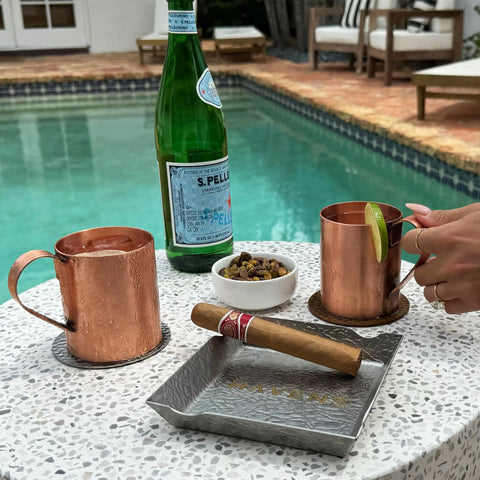 A Havens Luxury Metals poolside table displays two Pure Copper Mule Mugs (USA Made), San Pellegrino, pistachios, and a cigar on an ashtray. A hand holds a mug with lime garnish, the shimmering pool in the background.