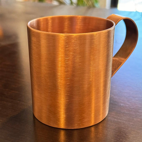 A Havens Luxury Metals Pure Copper Mule Mug, expertly handcrafted in the USA, features a smooth brushed finish and curved handle, resting on a dark wooden surface.