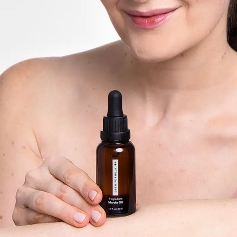 A woman with light skin smiles gently, holding an Open Formula Marula Oil brown dropper bottle (1 fl oz/30 ml) against her bare shoulder. This nourishing oil hydrates skin and protects hair. The background is plain white.