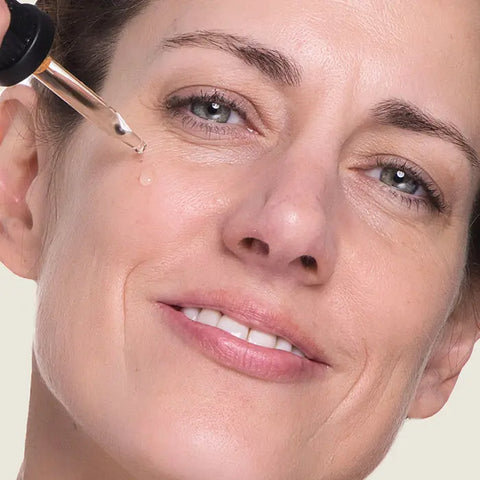 A woman smiles as she uses a glass dropper to apply Open Formula Marula Oil under her eye, deeply moisturizing her skin for a radiant look.