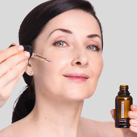 A woman with fair skin smiles softly as she moisturizes her cheek with Open Formula Marula Oil, using a dropper from a small brown bottle. Her dark hair is pulled back, set against a plain gray background.
