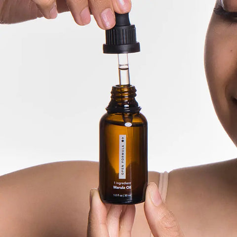 A person holds a dropper above an open amber bottle labeled Open Formula Marula Oil against a plain background, highlighting the oils hair-moisturizing and protective benefits, with part of their face and shoulder in view.