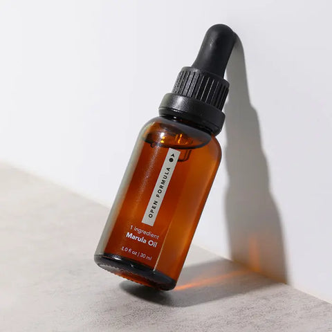 A brown glass dropper bottle of Open Formula Marula Oil (1.0 fl oz | 30 ml) with a black cap leans against a white surface; the single-ingredient oil naturally moisturizes and protects hair.