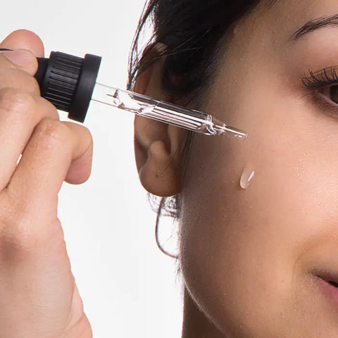 A close-up of a woman applying Open Formula Rosehip Oil to her cheek with a dropper, highlighting a droplet on her skin for radiant, natural skincare.