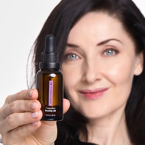 A dark-haired woman smiles while holding a brown glass dropper bottle labeled “Rosehip Oil” from Open Formula toward the camera, highlighting this natural skincare essential.