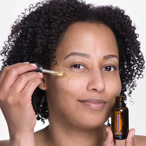 A person with curly hair uses a dropper to apply Open Formula Rosehip Oil to their cheek from a brown glass bottle, smiling slightly and enjoying their natural skincare routine.