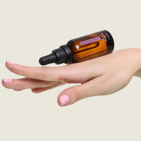 A hand with light pink nail polish balances an amber dropper bottle of Open Formula Rosehip Oil on the back of the fingers against a plain, light background, evoking a sense of natural skincare.